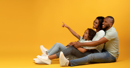 Joyful african american family sitting on floor, looking aside, cute girl pointing finger at copy...