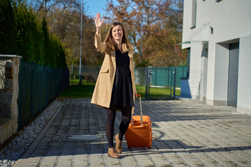 Woman pulling suitcase and waving hand near modern building