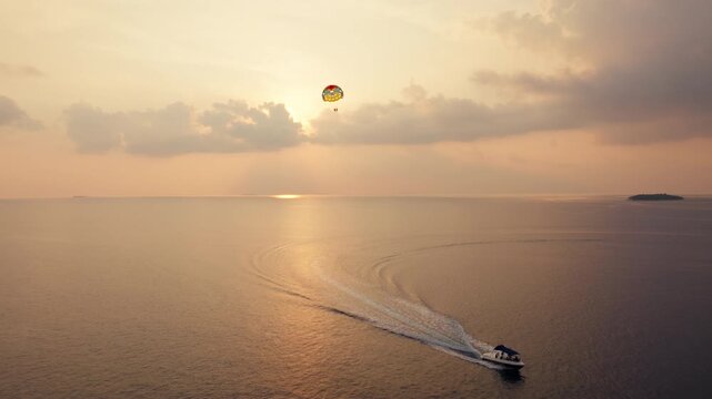 A stunning aerial view of a parasailer soaring high above the tranquil ocean as a motorboat cruises into the beautiful sunset. Experience freedom and adventure on this tropical escape.