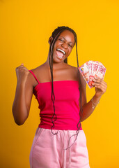 A Joyful young black woman with braided hair celebrates against a bright yellow background. She wears a pink top and pants, holding a fan of Ghanaian 200 Cedi notes while cheering with a raise fist