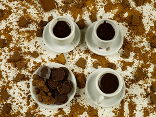 White cups with hot chocolate, a saucer with small pieces of chocolate, pieces of milk chocolate, cacao powder are on a white background, top view