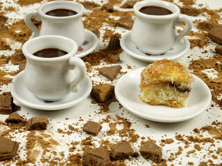 White cups with hot chocolate, a saucer with small piece of pie, pieces of milk chocolate, cacao powder are on a white background, side view
