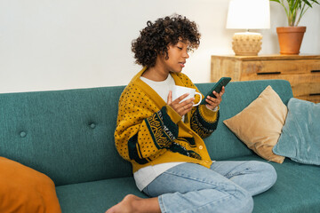Good morning. African girl drinking coffee holding smartphone sitting on couch at home Woman with...