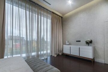 A stylish bedroom showcases wide windows draped with soft curtains, allowing natural light to...