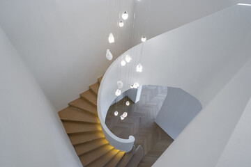 A spiral staircase with wooden steps curves upward. Hanging lights are positioned along the staircase. White walls surround the area, creating a spacious look.