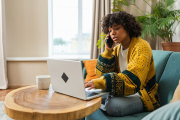 African American woman freelancer using laptop talking on phone at home office. Happy business girl having virtual meeting. Woman using phone laptop for communication working indoors. Remote work