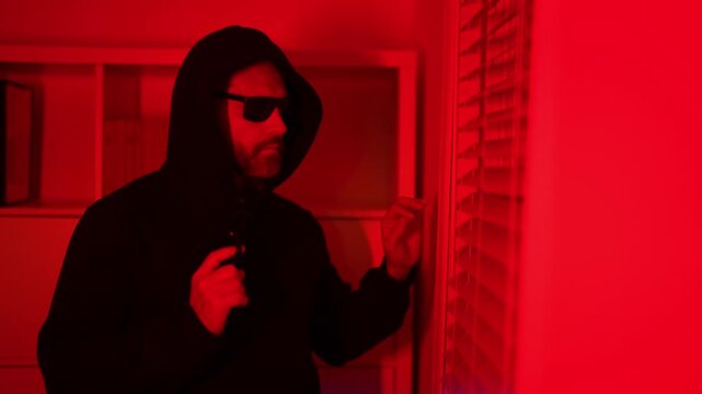 Burglar man breaking into house at night. Burglar near window. Criminal man attempting robbery indoors. Hooded burglar spying through window blinds. Burglar in home crime. Thief man.