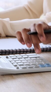 Vertical video of a person's hands using a calculator and writing notes in a notebook at a desk. Financial planning and budgeting concept.
