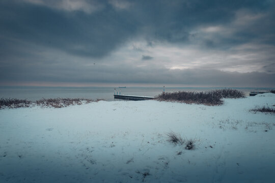 A scenic winter view from Raa Vallar in Helsingborg, Sweden. The image shows a snow-covered coastline and beach leading toward a calm sea with a wooden pier.