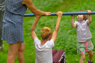 children practicing on horizontal bar with parent assistance, young boy holding bar while toddler reaches up, adult guiding hands, grassy park setting, casual summer clothing,