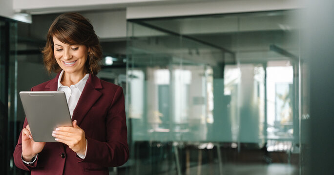 Businesswoman CEO holding digital tablet using fintech tab application standing at workplace in office. Latin mature adult professional business woman in suit looking at online trade app. Copy space