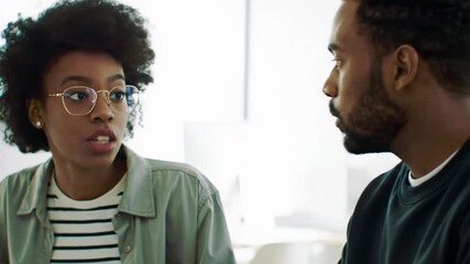 Young african american professionals in a sun-drenched office. Woman in sage green gestures, man intently observes. Blurred background, soft natural light, pan right subtly follows. Collaborative