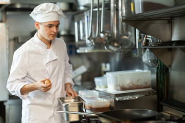 Diligent young male cook preparing dish on the stovetop in busy restaurant kitchen