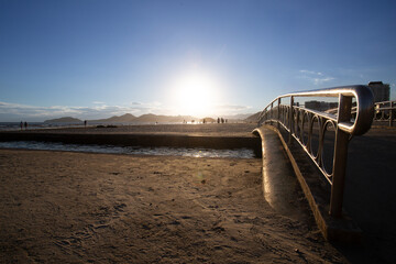 Golden sunset view at Canal 3 bridge on Santos beach, Brazil, with metallic railings, sand, sea, and silhouettes of people in the distance.