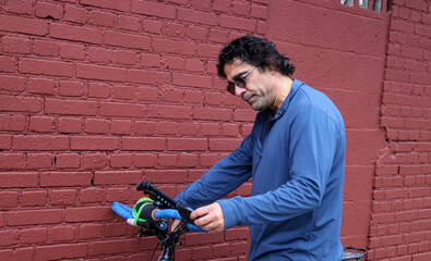 man in sunglasses checking on his bicycle (tire pressure bike maintenance) casual clothes commuter brick wall background urban cycling cyclist biker rider road mountain touring brakes handlebars nyc u