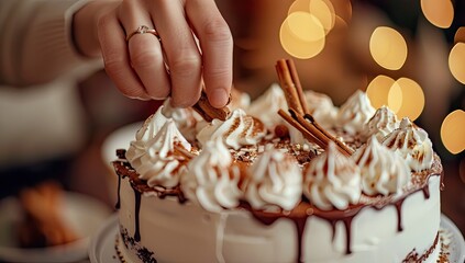 A hand carefully decorates a frosted cake with cinnamon sticks and whipped cream