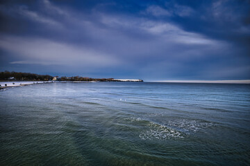 A dramatic winter seascape shows a wide, calm ocean with gentle waves rolling toward a quiet, snow-dusted coastline under a deep blue, stormy sky.