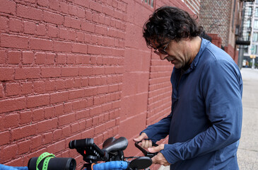 man in sunglasses checking on his bicycle (tire pressure bike maintenance) casual clothes commuter brick wall background urban cycling cyclist biker rider road mountain touring brakes handlebars nyc u