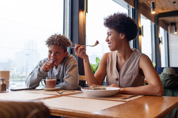 African American woman and her son are sitting in cafe drinking coffee and eating dessert, curly-haired teenager and his mother are having lunch in restaurant