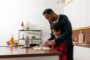 African American man and son cooking breakfast together in the kitchen, teenager cutting banana and cooking with father at home