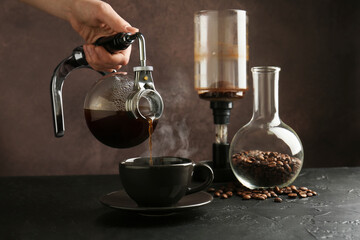 Woman pouring coffee into cup from siphon (vacuum) coffeemaker at black table, closeup