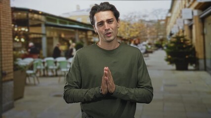 Man with hands pressed together in pleading gesture on street cafe walkway wearing green shirt and watch, palms visible; regret plea.