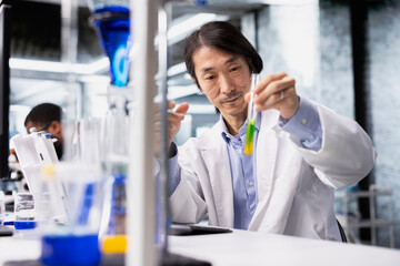 Old biochemist using laboratory glassware filled with green solution at workstation in research facility. Asian lab expert looking at cylinder with chemical fluid used for scientific experiments