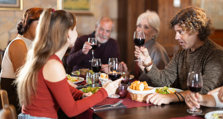 Large friendly family gathered at table to have lunch in restaurant. Relatives eat, drink wine, and socialize. People participate in discussion during meeting.