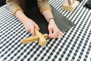Artisan using a wooden strap cutter to slice a strip of black leather on a checkered workbench.