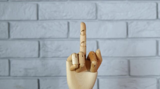 Wooden mannequin hand showing middle finger gesture. Articulated wooden hand slowly rising against a white brick wall background, showing an obscene and offensive gesture by raising the middle finger