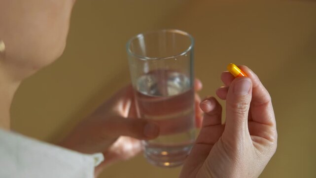 Woman taking medication pill with a glass of water. Unrecognizable woman taking a daily dose of medication, swallowing an orange and yellow capsule with a full glass of water for health, wellness