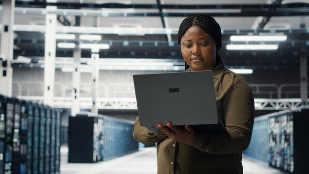 Data center workplace programmer using laptop to coordinate deployment of new server architecture. Server room worker using notebook to do rollout of cybersecurity framework, protecting data, camera B - Powered by Adobe