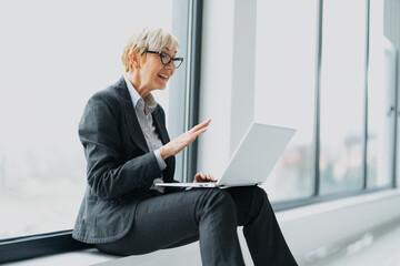 A professional middle-aged blonde businesswoman having a video call on her laptop in a bright office