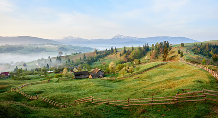 Tranquil morning landscape with lush green valley dotted with rustic farm buildings and winding wooden fences. Mist gently blankets lower areas, distant snow-capped mountains and pastel sky.