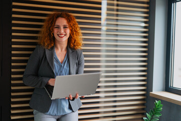 A young professional businesswoman with vibrant red hair standing in a modern office and holding a laptop