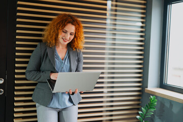 A young professional businesswoman with vibrant red hair standing in a modern office and holding a laptop