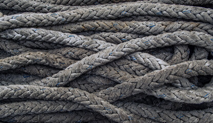 Close-up of weathered, grey nautical ropes tangled in a heap. The thick, braided texture shows detailed wear with subtle blue fibers and dark shadows