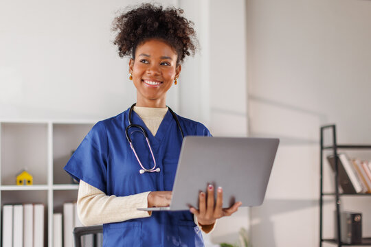 Medical professional in blue scrubs working on laptop, African american in afro hair smiling while standing in well-lit healthcare environment

