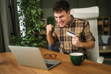 Young man celebrating successful online shopping using laptop and credit card