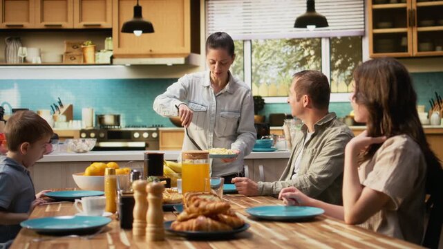 Family sharing scrambled eggs and pastry for breakfast at home, parents serving omelettes and toast with little boy and pre-teen girl. Warmth, relaxation and joyful togetherness. Camera A.