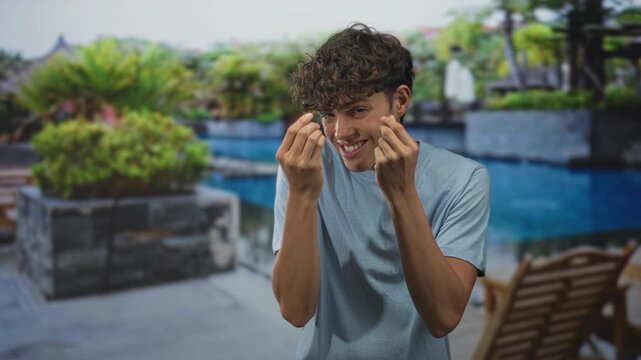 Teenage boy smiling and making money gesture with fingers near face by pool at building terrace, curly hair and light t shirt; playful invitation.
