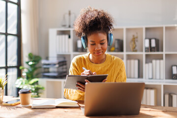 African american in afro hairwearing headphones using laptop in cafe, writing notes, attractive female student learning language, watching online webinar, listening audio course, e-learning education 