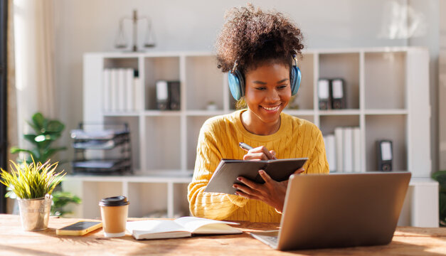 African american in afro hairwearing headphones using laptop in cafe, writing notes, attractive female student learning language, watching online webinar, listening audio course, e-learning education 