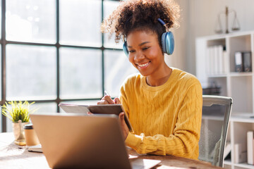 African american in afro hairwearing headphones using laptop in cafe, writing notes, attractive female student learning language, watching online webinar, listening audio course, e-learning education 