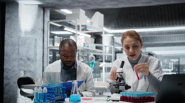 Pathologist examining biopsy slide while colleague documents diagnostic findings on clipboard. Medical experts working together to identify petri dish disease markers in clinical lab, camera A