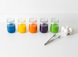Jars with colorful egg dye liquid, white eggs on white surface, metal tongs. Preparation for Easter decoration concept. Children craft activity.