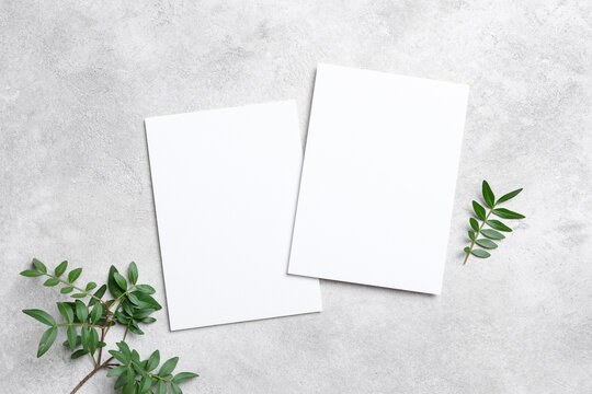 Blank invitation card mockup with two empty cards on a textured surface surrounded by green foliage, ideal for customizable designs and creative projects with copy space