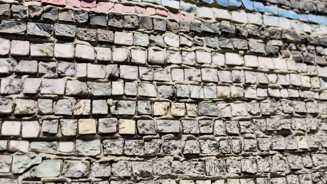 Vibrant stone mosaic wall closeup showing green and blue square tiles set in rough mortar, weathered grout lines, uneven beige stones, sunlit texture, urban exterior, decorative.