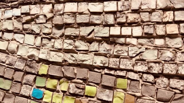 Vibrant stone mosaic wall closeup showing green and blue square tiles set in rough mortar, weathered grout lines, uneven beige stones, sunlit texture, urban exterior, decorative.