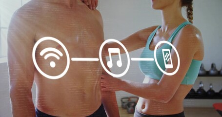 Adjusting trainer in teal bra pressing shirtless man torso in gym, showing Wi?Fi music phone icons